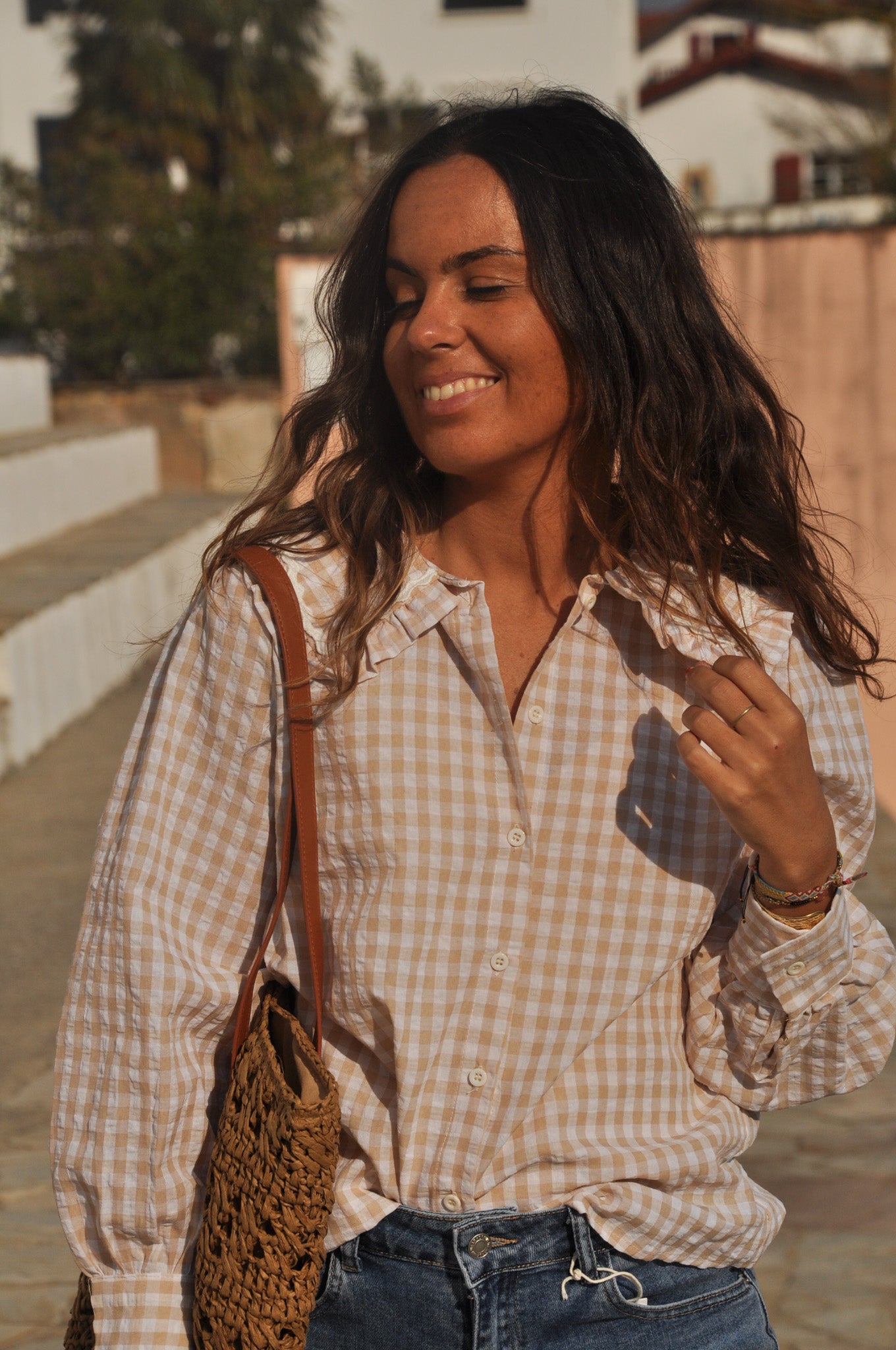 Femme souriante portant une chemise à carreaux vichy beige et blanc avec col volanté et jean bleu.