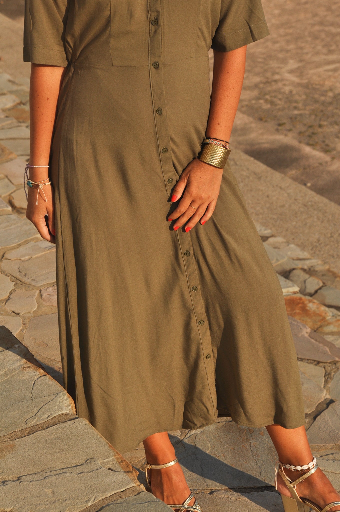 Femme portant une robe longue kaki fluide avec sandales dorées en extérieur.