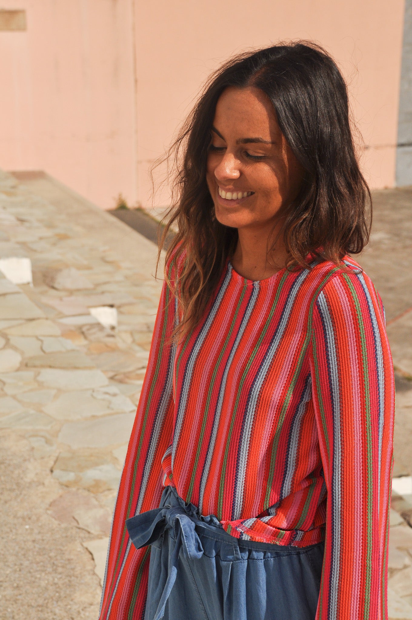 Femme souriante portant un pull léger à rayures verticales multicolores (rouge, corail, rose, vert, gris bleu) et un jean
