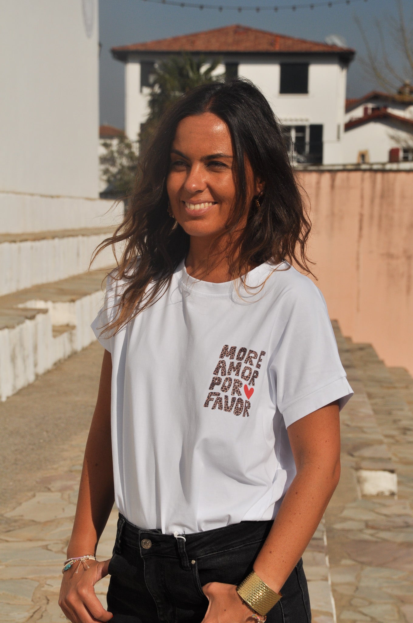 Femme souriante portant un t-shirt blanc 'More Amor Por Favor' imprimé léopard et un jean noir en extérieur.